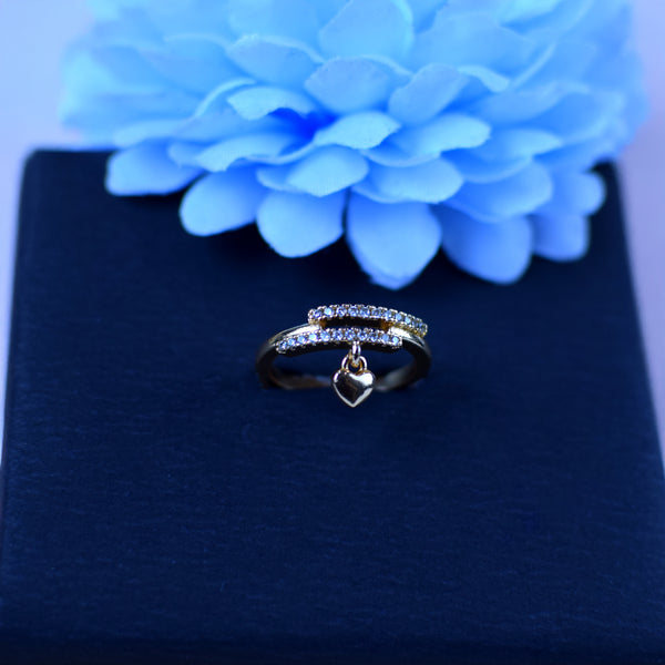 Gold ring with heart charm on a dark surface with a blue flower in the background